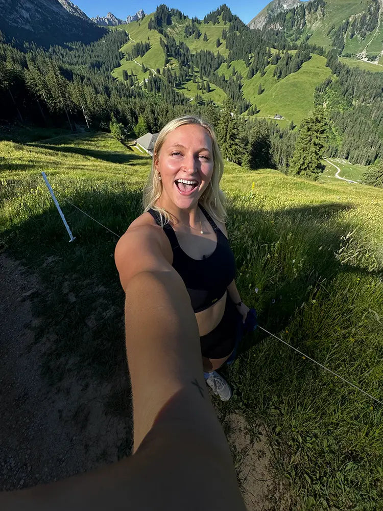 Woman smiling in black sports bra on mountain trail with green hills behind.