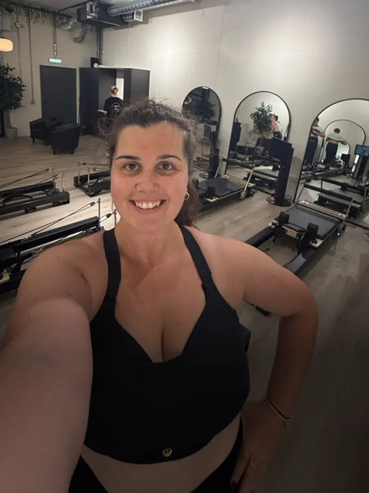 Woman in black PEBE sports bra taking a selfie in a reformer pilates studio.