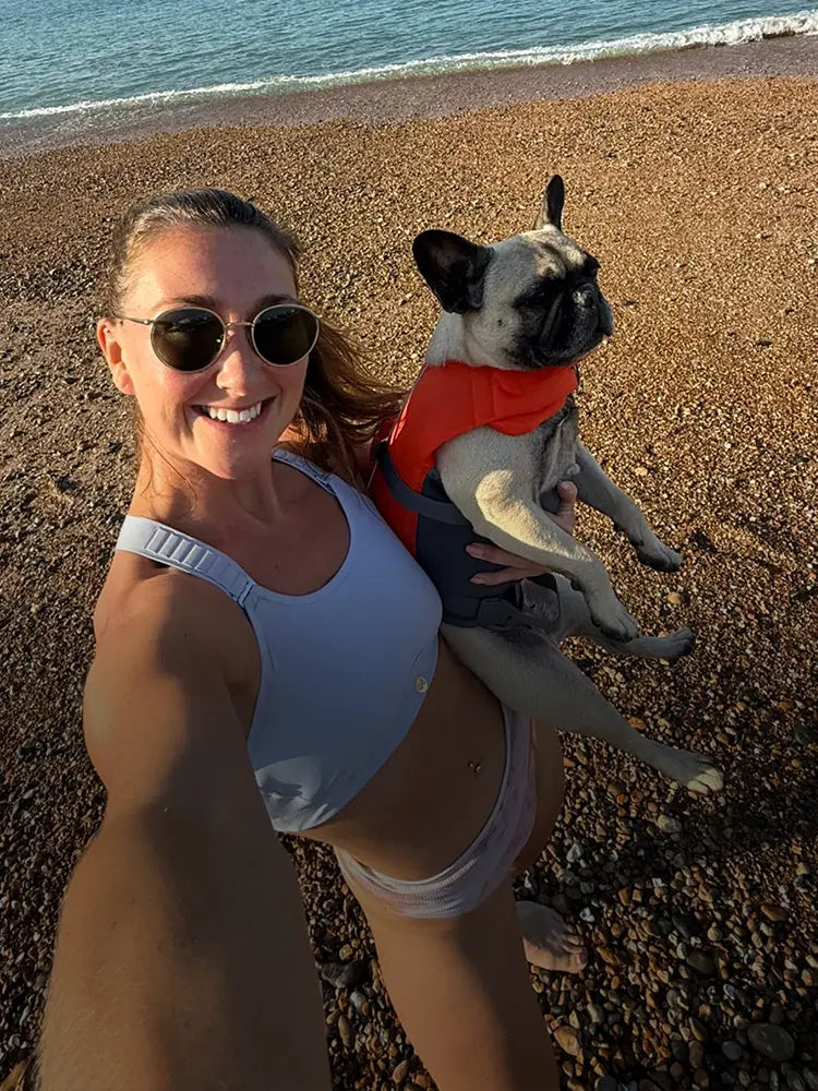 Woman in blue sports bra holding dog on pebble beach by the sea.