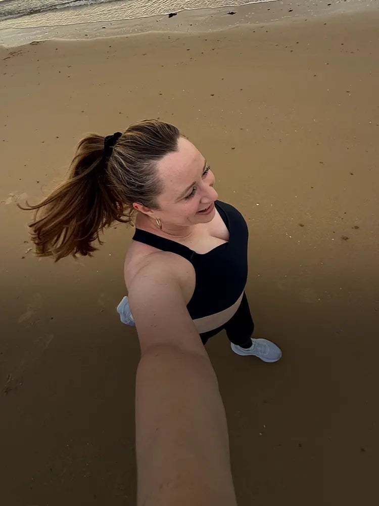 Woman jogging on beach in black sports bra with ponytail and white trainers.