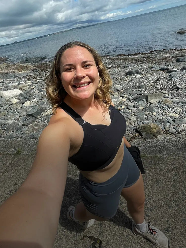 Woman smiling in black sports bra by the sea on a rocky coastal path.